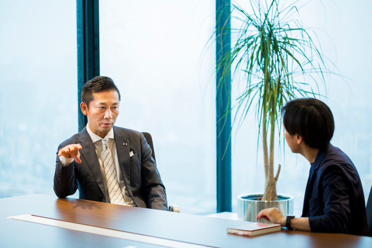 奥には手振りを交えて説明する島田社長。手前には話を聞く青野。