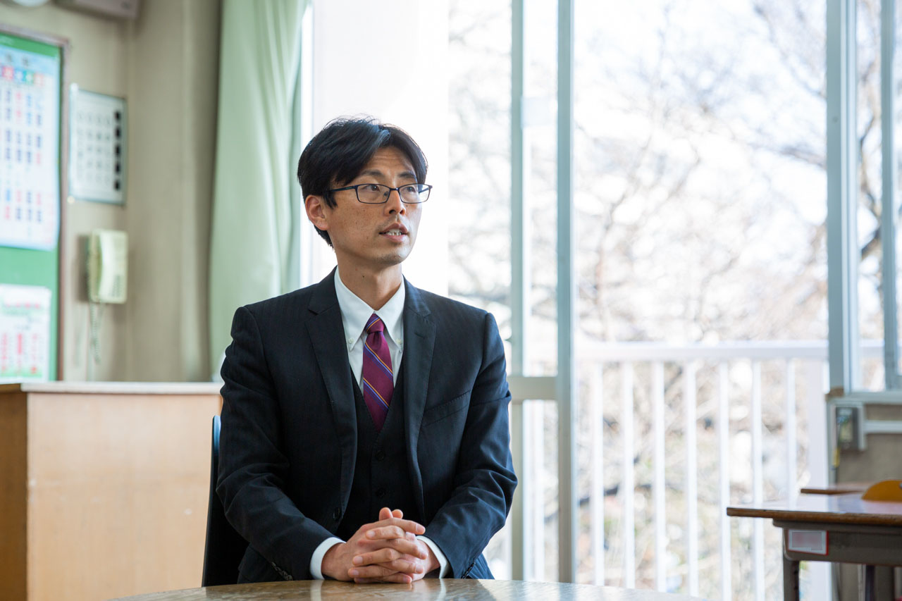 教室で椅子に腰かけて話す森田先生。背景には花咲く桜が窓越しに見えている